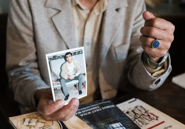 A close-up of a hand holding a color photograph of a young man seated on a cot.