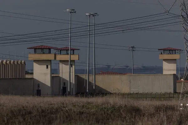 Three guard towers with red roofs rise above a very high wall. Multiple power lines crisscross the sky.