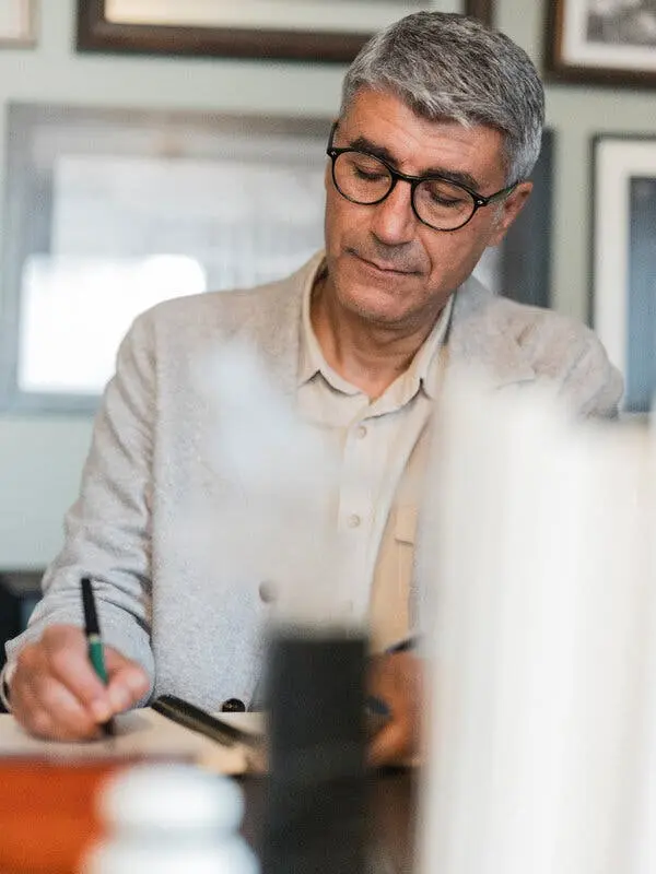 Mr. Çomak seen from the waist up seated at a desk. He is writing with a pen held in his right hand.
